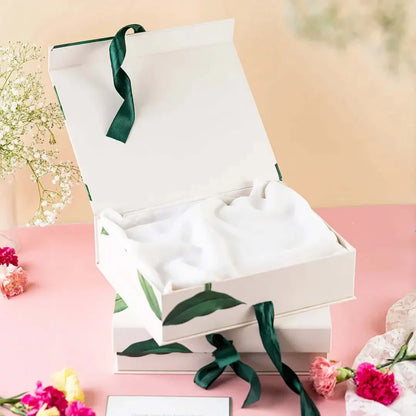 Open gift box with white tissue paper and green ribbon on a pink surface with flowers.