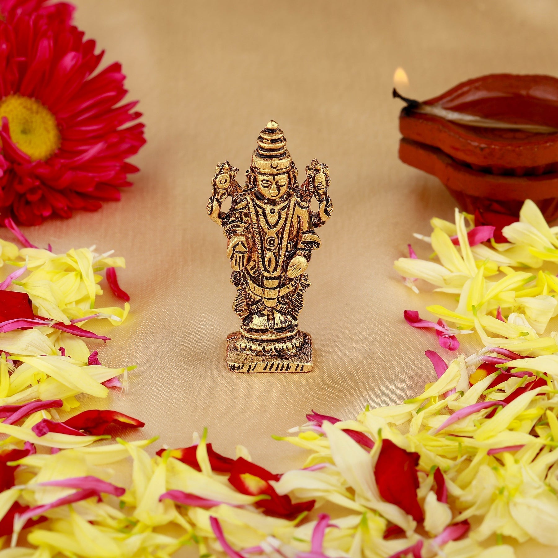 Decorative statue of a deity surrounded by flowers and a lit lamp on a beige background