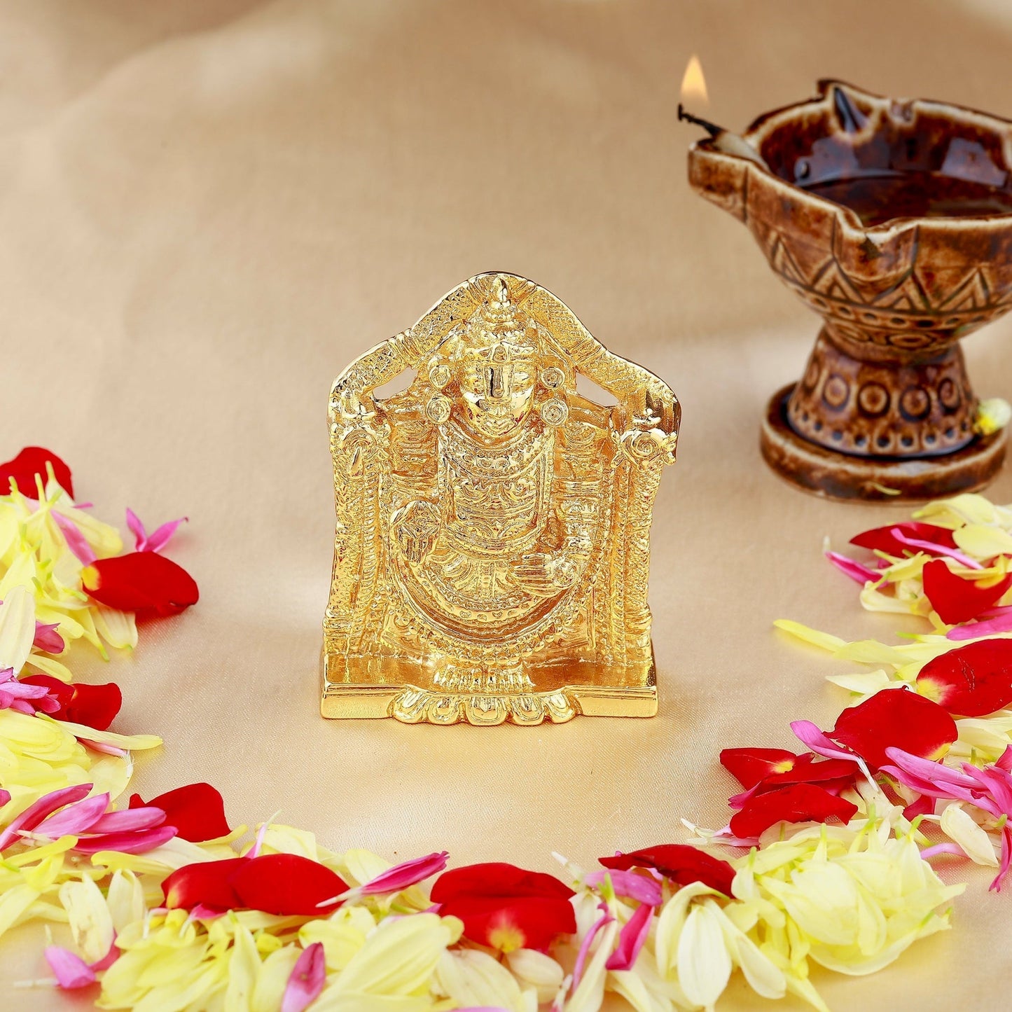 Gold-colored deity statue with flowers and a lit oil lamp on a beige background