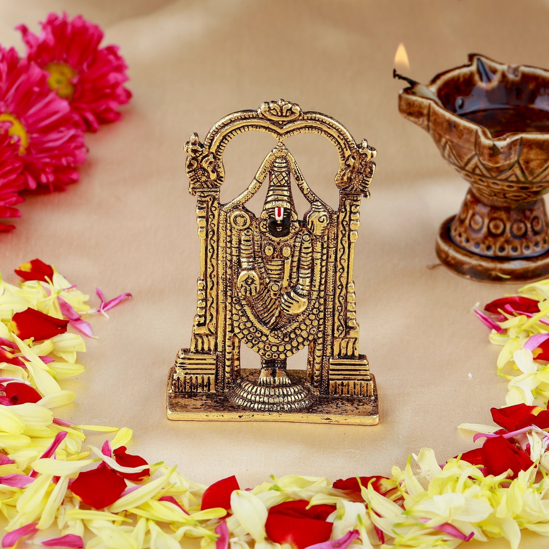 Bronze idol of a deity surrounded by flowers and a lit oil lamp on a light background