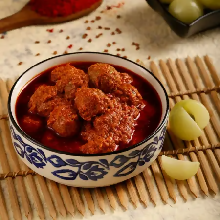 Bowl of meatballs in a red sauce on a wooden mat with a spoon and grapes in the background.
