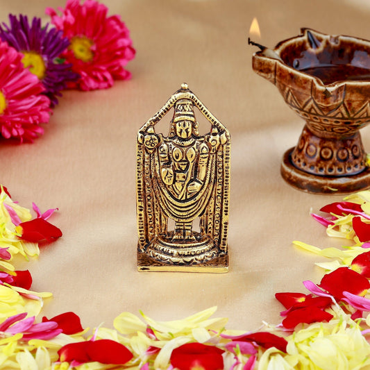 Bronze idol of a deity surrounded by flowers and a lit oil lamp on a beige background