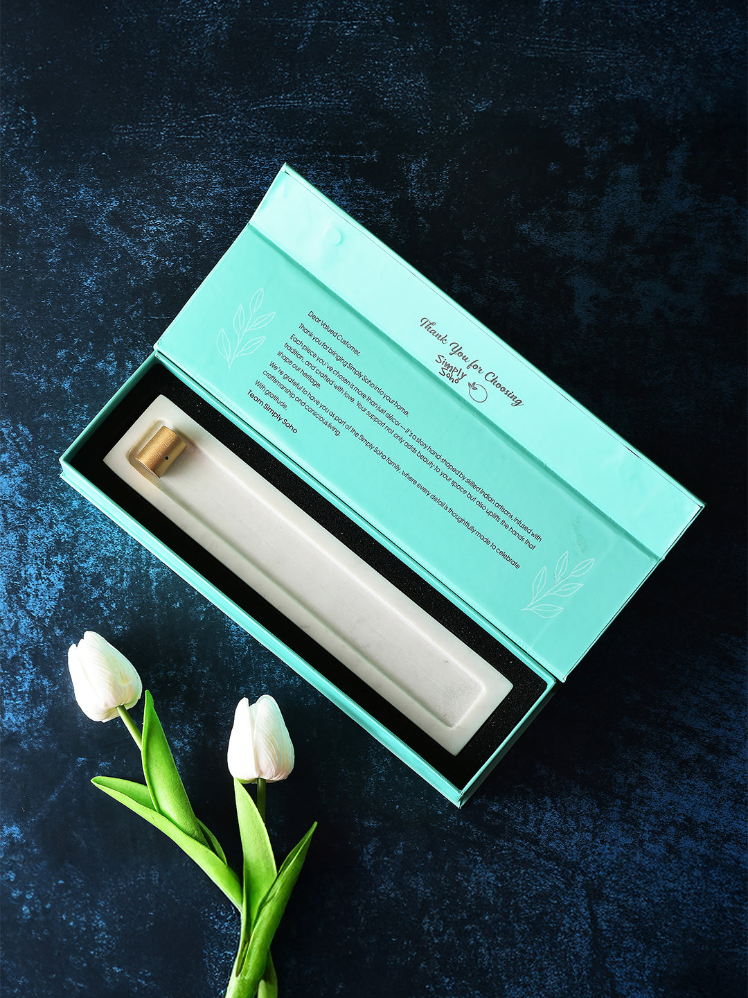 White ceramic object in a teal box on a dark textured surface with white tulips.