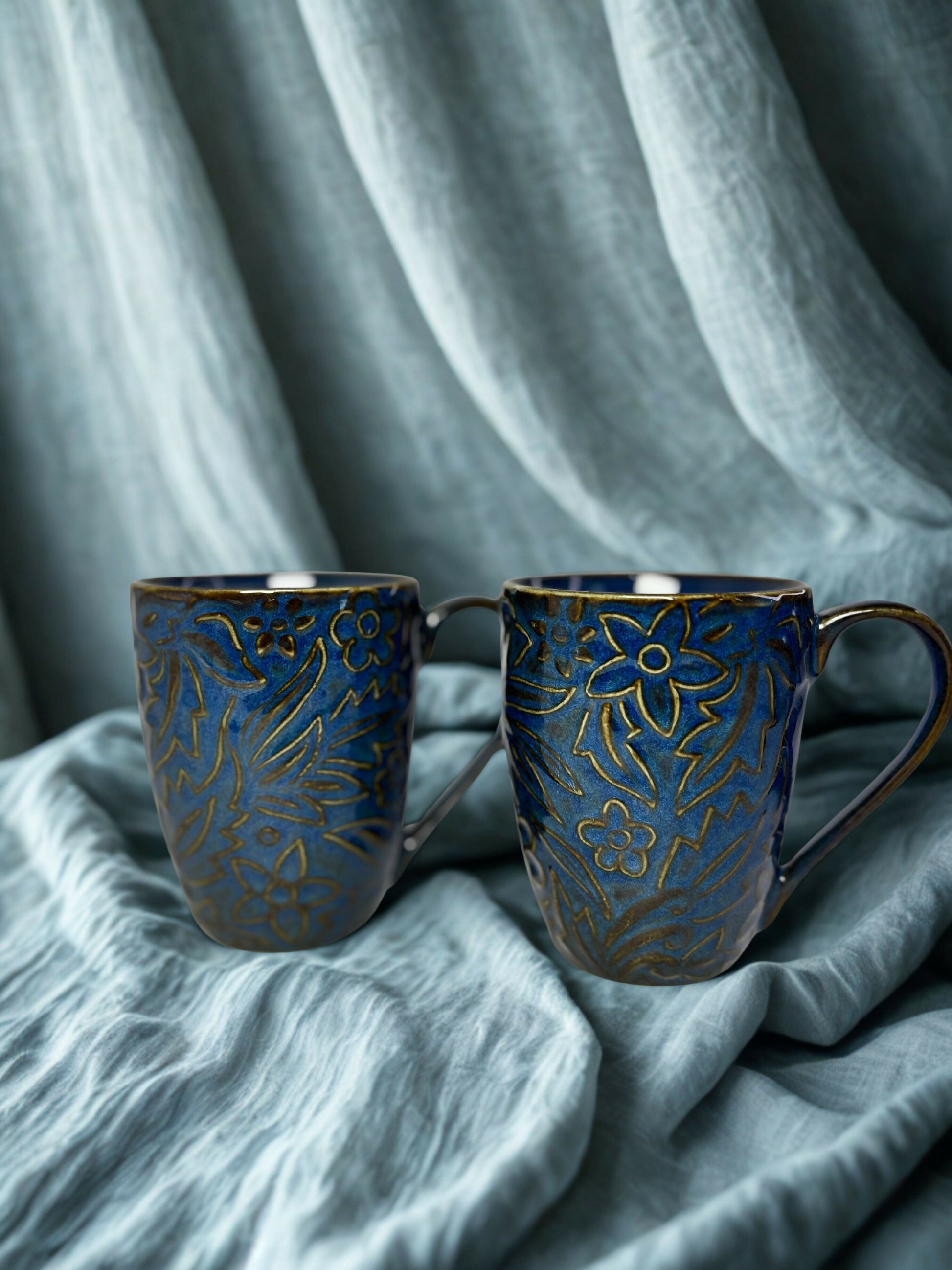 Two blue ceramic mugs with gold patterns on a textured gray fabric background