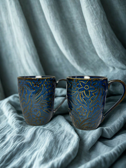 Two blue ceramic mugs with gold patterns on a textured gray fabric background