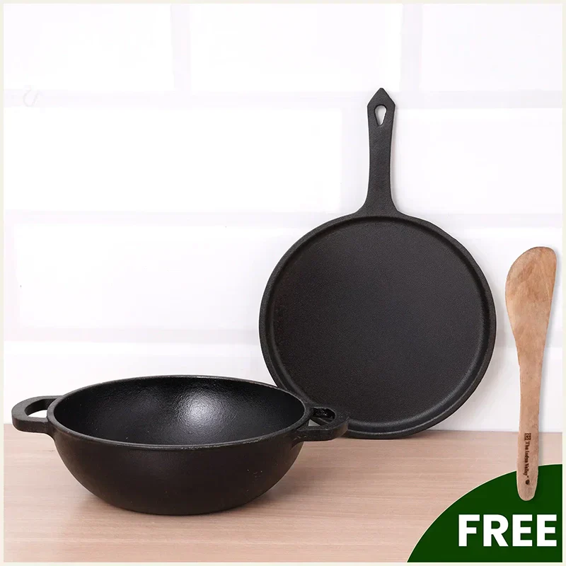 Black cast iron wok and skillet on a light wooden surface with a wooden spatula, against a white background.