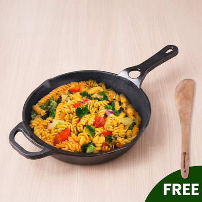 Black cast iron skillet with pasta and vegetables on a light wooden surface, accompanied by a wooden spoon.