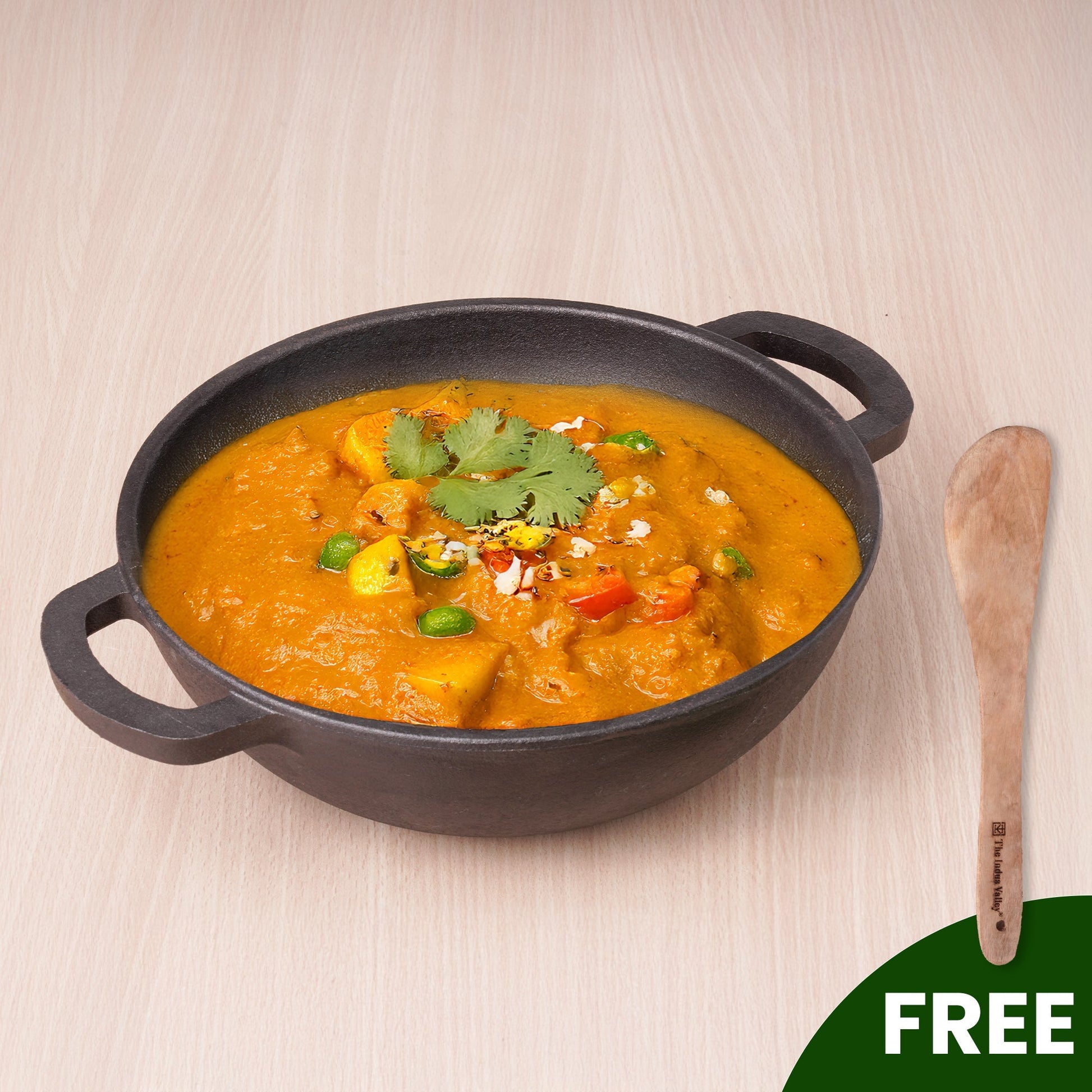 Black cast iron skillet with orange curry and garnishes on a light wooden surface, with a wooden spoon to the side.