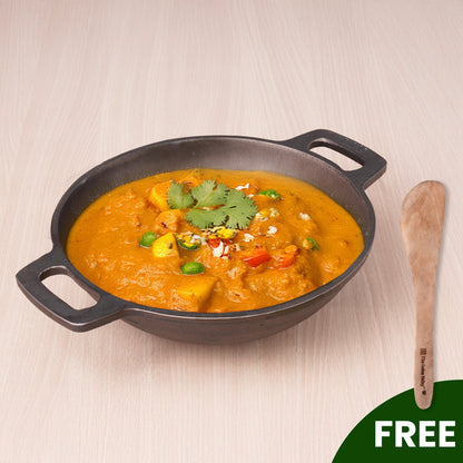 Black skillet with orange curry dish on a light wooden surface, featuring a wooden spatula.
